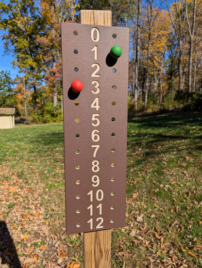 Brown ColorCore bocce scoreboard with tan numbers from 0 to 12 and red and green scoring pegs, mounted outdoors on a wooden post.