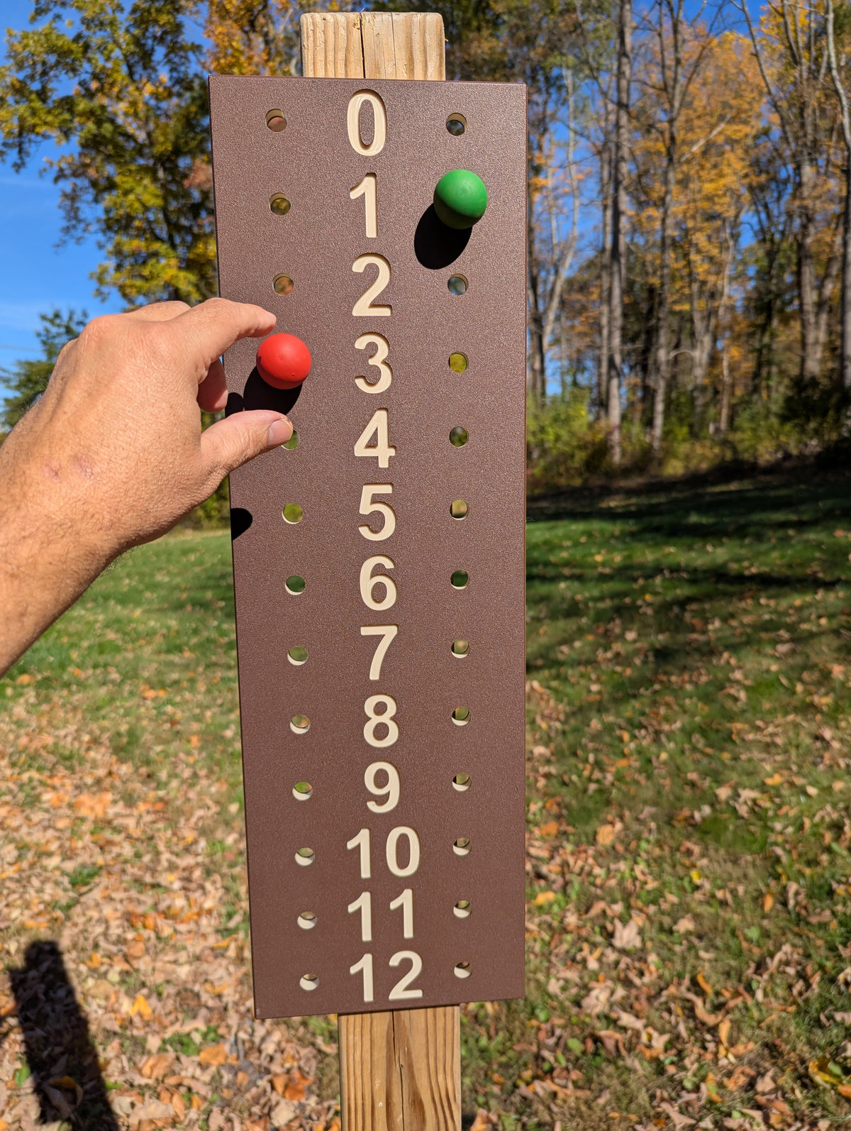 Hand adjusting a red scoring peg on a brown bocce scoreboard with tan numbers, mounted on a wooden post.