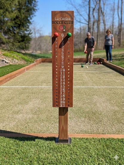 A wooden lawn game scoreboard labeled 'Le Club du Bocce' stands on grass in an outdoor backyard setting with a bocce court behind it and two people standing in the distance.