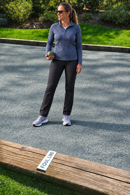 A woman wearing a slate blue long-sleeve quarter-zip pullover and dark gray athletic pants stands on an outdoor bocce court, holding a bocce ball 